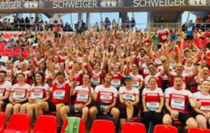 Eine Menge fröhlicher Freiburg-Anhänger in rot-weißer Kleidung, viele heben ihre Hände, sitzen bei einer Sportveranstaltung auf roten Stadionsitzen.