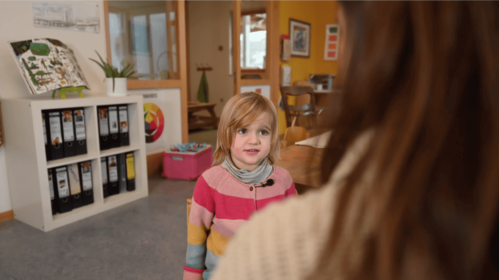 Ein kleines Kind in einem bunt gestreiften Pullover unterhält sich mit einem Erwachsenen in einem gemütlichen Raum voller Spielsachen und Bastelutensilien in Freiburg. Das Kind sieht aufmerksam und neugierig aus.