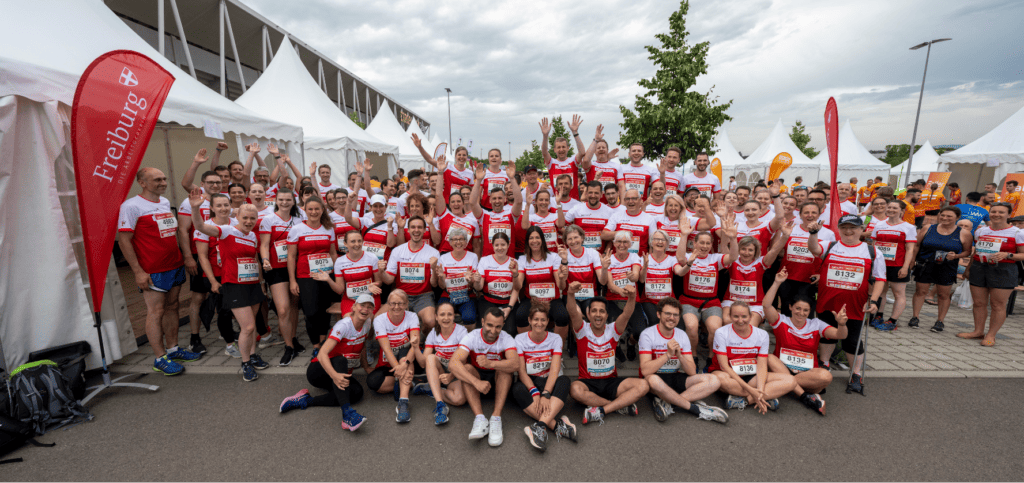 Eine große Gruppe Läufer in rot-weißen Uniformen posiert fröhlich gemeinsam bei einer Laufveranstaltung in Freiburg, im Hintergrund sind Veranstaltungszelte und andere Teilnehmer zu sehen.