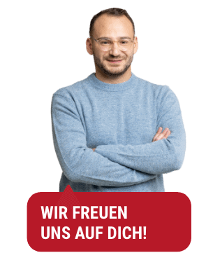 Ein Mann mit Brille und blauem Pullover steht mit verschränkten Armen da und lächelt. Unter ihm steht auf rotem Hintergrund ein deutscher Text mit der Aufschrift „Wir freuen uns auf ein Treffen mit Ihnen in Freiburg!“.