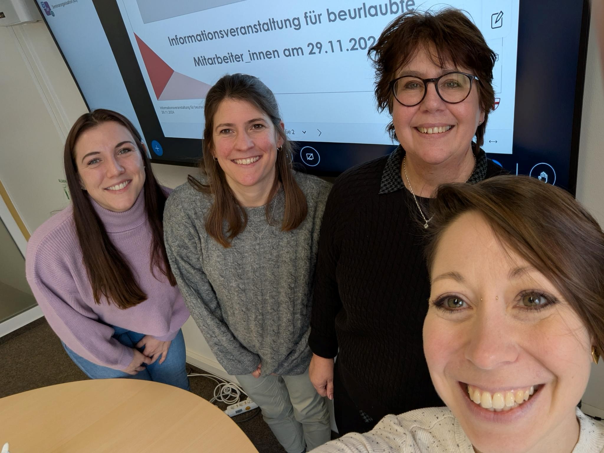 Vier Frauen sind auf einem Selfie zu sehen. Alle lachen. Im Hintergrund läuft eine Präsentation, die die Teilnehmenden über das Thema der Veranstaltung informiert.