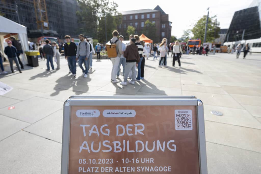 Auf dem Schild zum „Tag der Ausbildung“ in Freiburg sind Veranstaltungsdetails mit QR-Code zu sehen. Im Hintergrund sind Gruppen von Menschen zu sehen, die sich Stände auf einem sonnigen Platz im Freien ansehen.