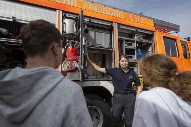 An einem sonnigen Tag demonstriert eine Feuerwehrfrau in Freiburg einer Gruppe aufmerksamer Teenager die Ausrüstung eines Feuerwehrautos.