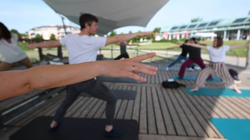 In Freiburg findet gerade ein Yoga-Kurs im Freien statt. Die Aufnahme erfolgt aus einem Winkel, der auf den ausgestreckten Arm einer Frau mit einem Ring fokussiert ist. Im Hintergrund sind unscharf andere Teilnehmer in Yoga-Posen zu sehen.