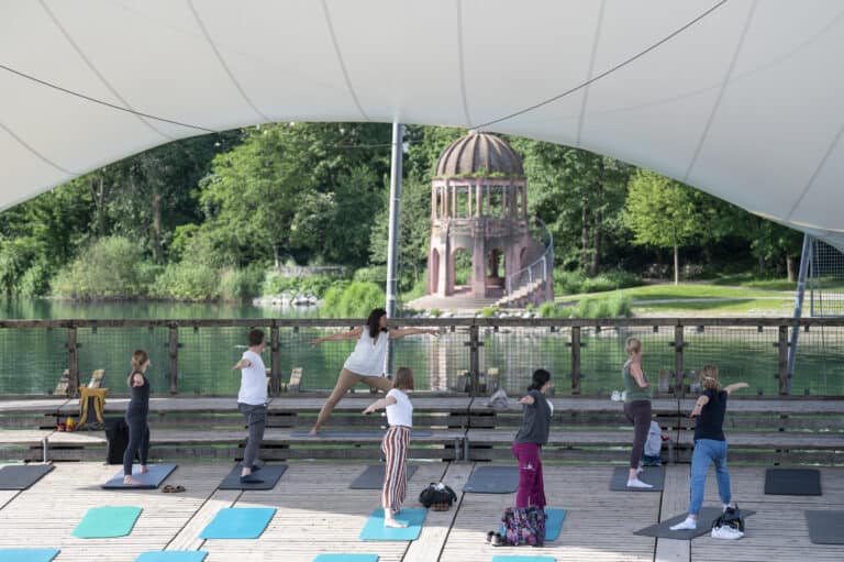 Auf einer Holzplattform an einem See in Freiburg findet unter einem großen weißen Baldachin eine Yogastunde statt. Menschen unterschiedlichen Alters führen Posen aus, im Hintergrund ein Pavillon und Bäume.