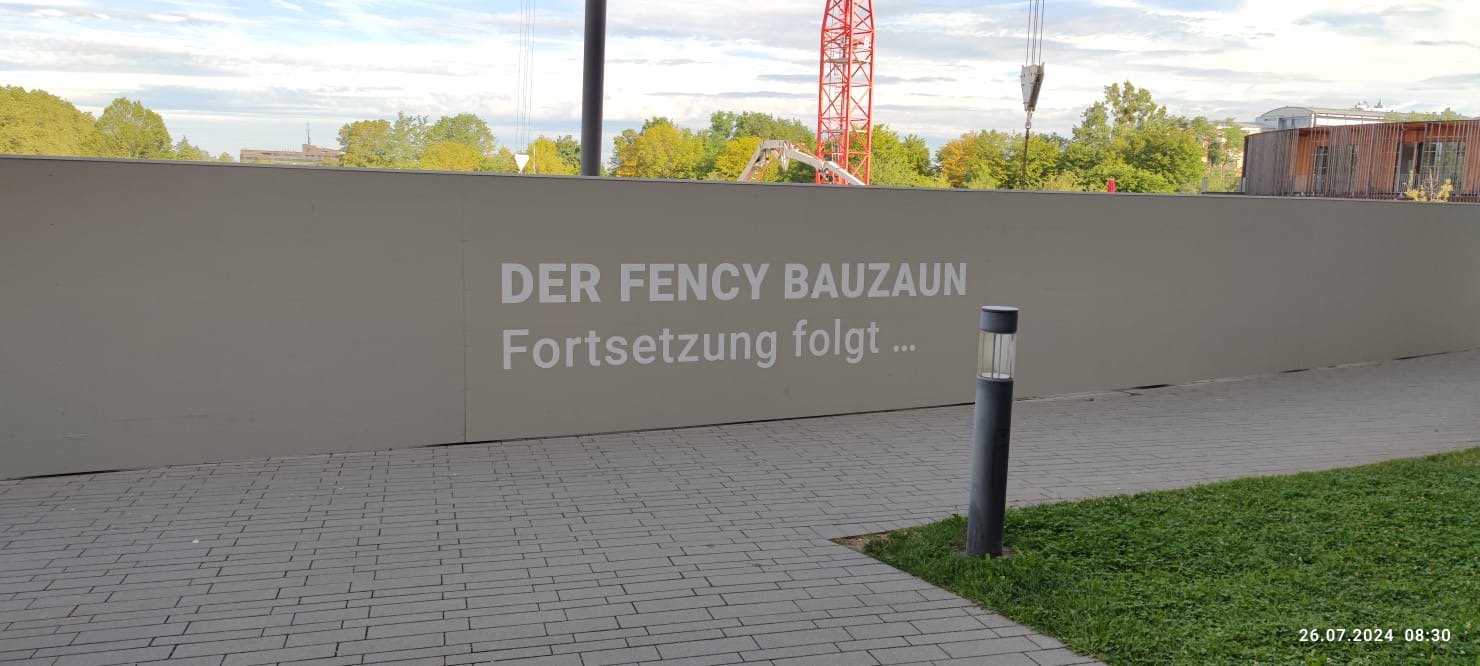 Ein grau bemalter Bauzaun aus Holz. Auf diesem stehen die Worte: "Der Fency Bauzaun. Fortsetzung folgt..."